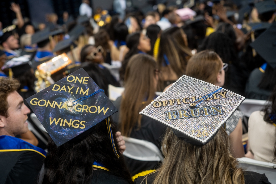 Two students wearing graduation caps with one reading "Day one, I'm gonna grow wings," and the other reading "officially a Bruin"