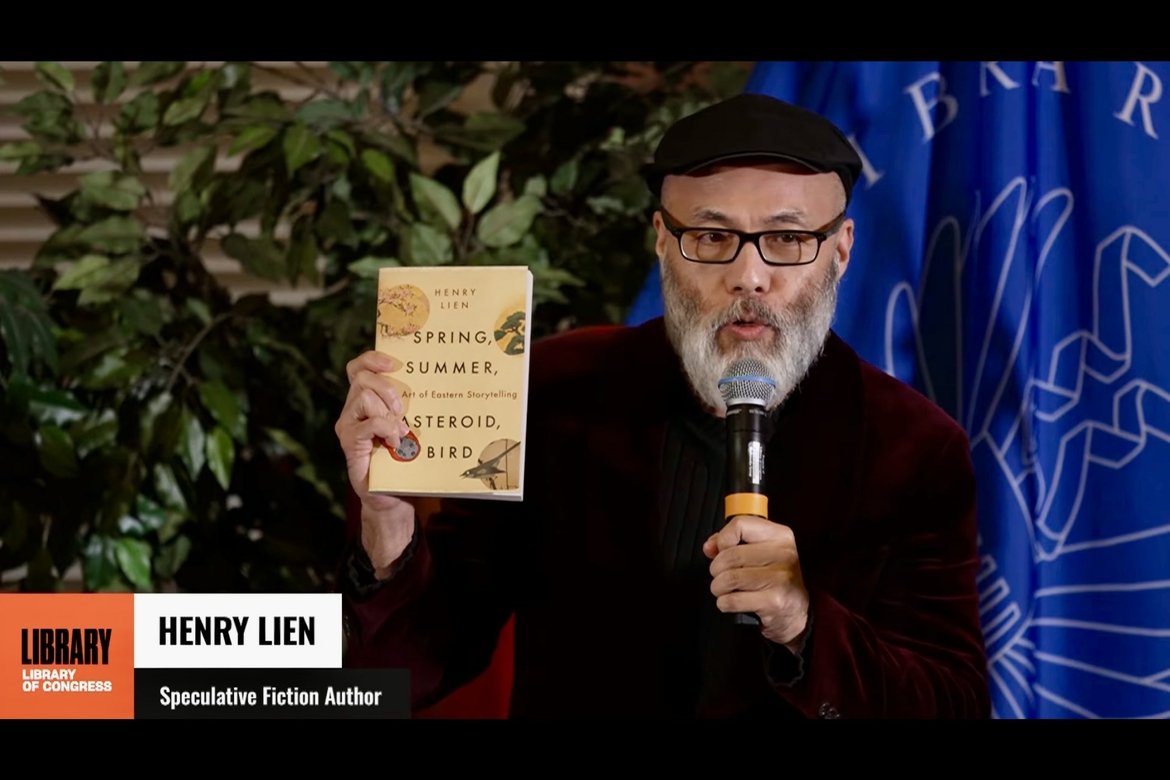 Henry Lien holds up his book while speaking in a microphone at the Library of Congress
