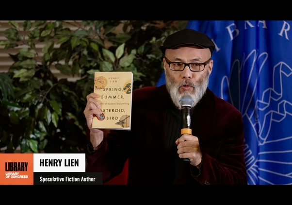 Henry Lien holds up his book while speaking in a microphone at the Library of Congress