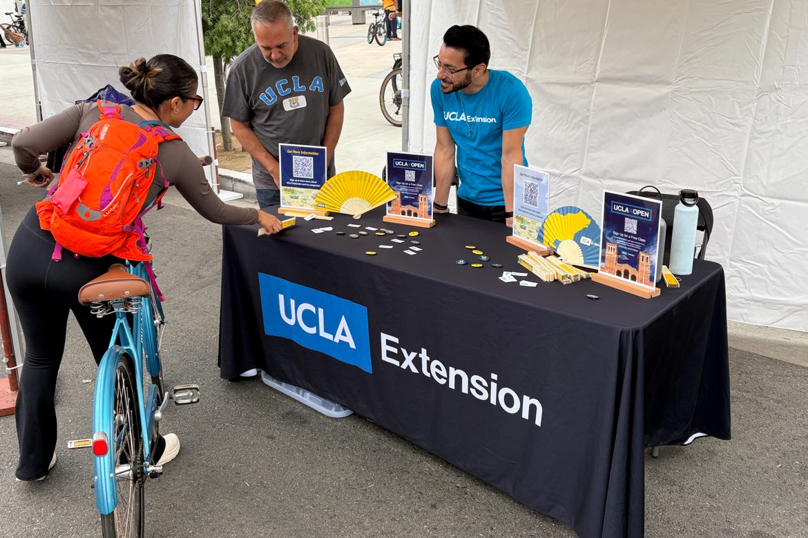A woman on a bicycle picks up a fan from a UCLA Extension table with two staff members