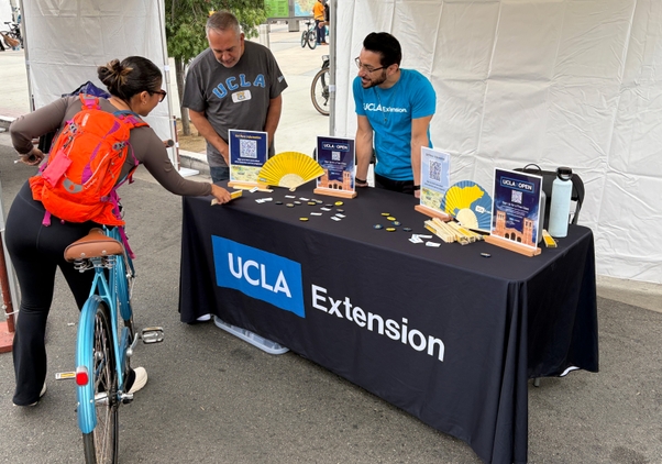 A woman on a bicycle picks up a fan from a UCLA Extension table with two staff members