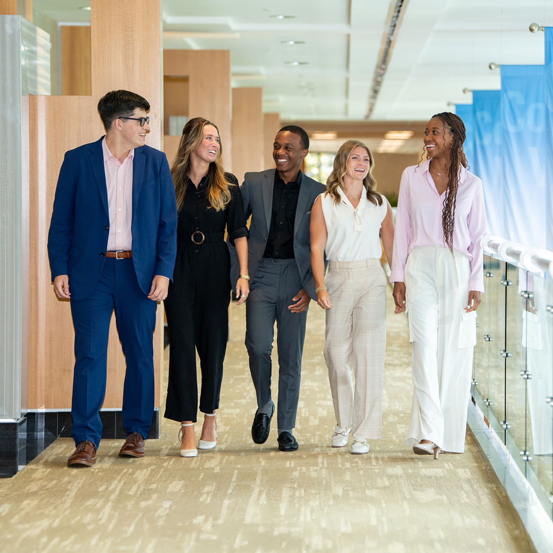 Five people in business attire walking and smiling in a modern corridor.